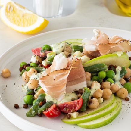 Insalata di legumi con mela Granny Smith e Speck Alto Adige IGP su piatto bianco, con limone sullo sfondo.