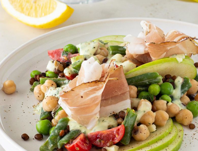 Insalata di legumi con mela Granny Smith e Speck Alto Adige IGP su piatto bianco, con limone sullo sfondo.