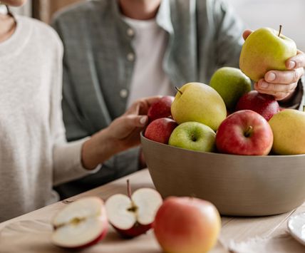 Manzana cortada sobre tabla, al fondo cuenco con manzanas, dos personas y manos que toman una manzana