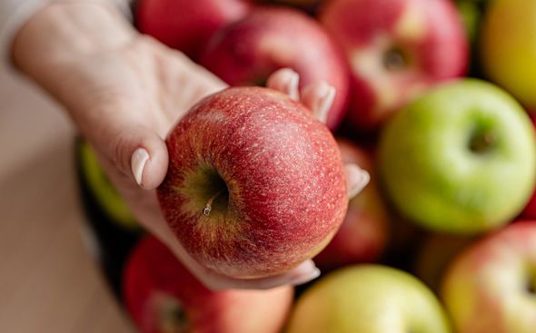 Primer plano de una manzana roja en una mano, al fondo borroso un cesto con manzanas frescas
