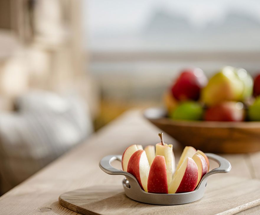 Manzana cortada sobre tabla de madera en primer plano, cuenco con manzanas variadas y montañas al fondo