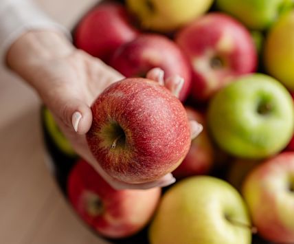 Manzana roja sostenida en la mano, detrás cuenco con manzanas de distintas variedades