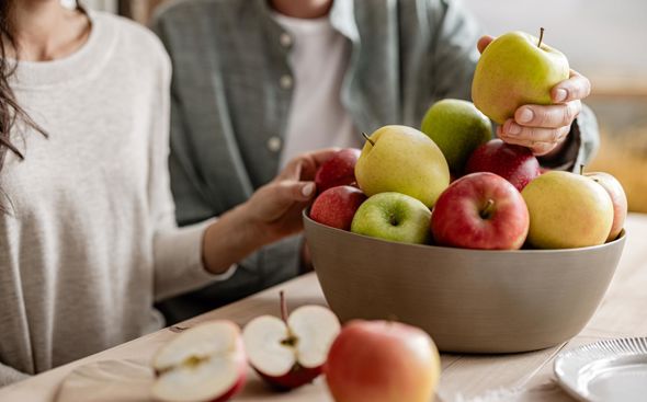 Mujer y hombre sentados en mesa de madera, ambos con una manzana en la mano; delante cuenco con manzanas variadas y manzana cortada sobre tabla de madera