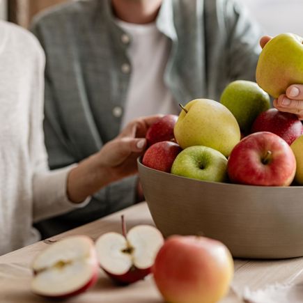 Donna e uomo seduti a un tavolo di legno, entrambi con una mela in mano; davanti ciotola di mele miste e mela tagliata su tagliere