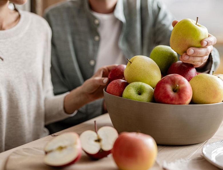 Donna e uomo seduti a un tavolo di legno, entrambi con una mela in mano; davanti ciotola di mele miste e mela tagliata su tagliere