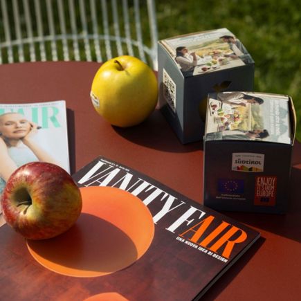 Due riviste Vanity Fair, due mele e due confezioni di cibo sono disposte su un tavolo rotondo rosso all'aperto, con sfondo erboso.