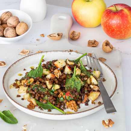Insalata di lenticchie croccanti, mele, noci e spinacino su piatto bianco con forchetta, con mele fresche, noci e gusci sullo sfondo.