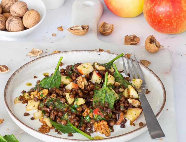 Insalata di lenticchie croccanti, mele, noci e spinacino su piatto bianco con forchetta, con mele fresche, noci e gusci sullo sfondo.