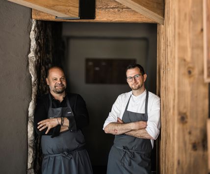 Dos hombres con delantales de cocina apoyados en paredes de madera y piedra